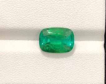 Puede incluir: Una vibrante esmeralda de talla cojín. La gema es de un color verde intenso y profundo con inclusiones internas visibles. La esmeralda está sobre un fondo blanco.