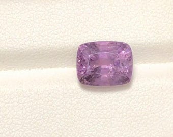 May include: A cushion-cut, light purple gemstone. The gem is set against a white background. The stone has a square shape with rounded corners and a faceted surface, reflecting light. The color is a soft, lavender hue.