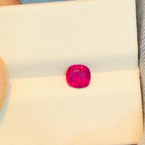 May include: A vibrant, cushion-cut, hot pink gemstone. The gem is set against a white background, showcasing its brilliant color and shape. The gemstone appears to be a ruby or similar precious stone.