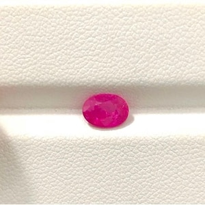 May include: A vibrant, oval-cut ruby gemstone, displaying a rich, deep pink hue. The gem is set against a white, textured surface, highlighting its brilliance. The ruby's facets catch the light, creating a sparkling effect. A close-up shot emphasizes the gem's clarity and color.
