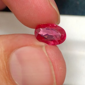 May include: A vibrant red, oval-cut gemstone, held between fingers. The facets of the gem catch the light, creating internal reflections. The polished surface suggests a ruby or similar precious stone, likely for jewellery.