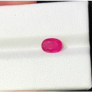 Puede incluir: Una gema de rubí de corte ovalado. El rubí muestra un rico tono rojo intenso, con características internas visibles. La gema se presenta sobre un fondo blanco, resaltando su color y claridad. Adecuado para la creación de joyas.