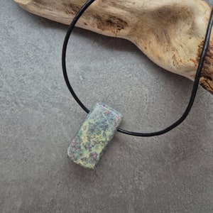 Op de afbeelding: Een groene en roze edelsteen hanger aan een zwart lederen koordsnoer. De hanger is rechthoekig en heeft een gevlekt patroon.