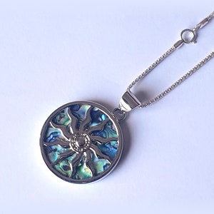Op de afbeelding: Een zilveren ketting met een ronde hanger met een blauw, groen en iriserend abalone-schelp zonontwerp.
