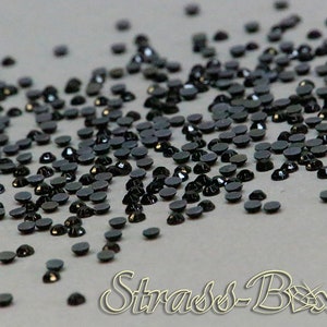 May include: A scattering of black rhinestones with a flat back and a faceted top. The rhinestones are on a grey surface.