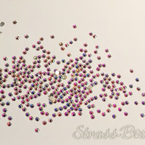 May include: A scattering of iridescent clear rhinestones on a white background. The rhinestones are small and have a rainbow effect.