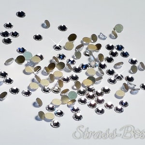 May include: A scattering of clear crystal rhinestones on a white background. The rhinestones are various sizes and shapes, with some having a silver backing.