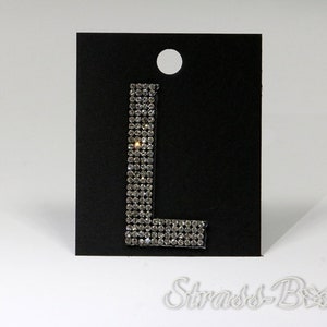 Könnte beinhalten: Ein silberner Strass-Buchstabe L auf schwarzem Hintergrund. Der Buchstabe besteht aus kleinen, klaren Strasssteinen, die in einem Gittermuster angeordnet sind. Der Buchstabe ist etwa 5 cm hoch und 2,5 cm breit.