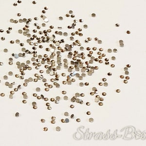 May include: A collection of small, circular rhinestones in shades of brown and silver, scattered on a white background. The rhinestones have a metallic appearance and reflect light. The image shows a close-up view of the scattered embellishments, creating a textured effect.