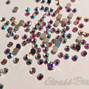 May include: A scattering of iridescent rainbow rhinestones on a white background. The rhinestones are various sizes and shapes, with some having a flat back and others having a pointed back.