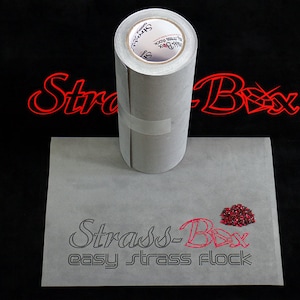 Puede incluir: Un rollo de tela gris de respaldo adhesivo con el texto "Strass-Box easy strass flock" impreso en él. Un pequeño montón de piedras de cristal rojas está al lado del texto.