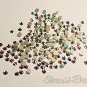 May include: A large quantity of iridescent, clear glass rhinestones. The rhinestones are scattered on a white surface.