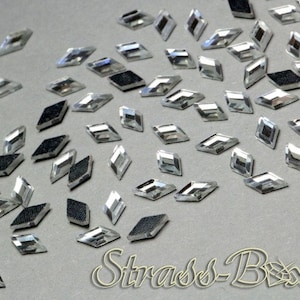 Op de afbeelding: Een verzameling heldere, ruitvormige strass-steentjes met een reflecterend oppervlak en een zwarte, getextureerde achterkant. De strass-steentjes liggen verspreid op een grijze ondergrond, met de tekst "Strass-Box" rechtsonder.