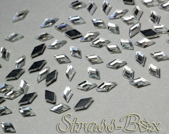 Puede incluir: Un conjunto de brillantes transparentes en forma de diamante, con una superficie reflectante y un reverso negro texturizado. Los brillantes están dispersos sobre una superficie gris, con el texto "Strass-Box" en la esquina inferior derecha.