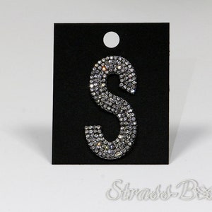 May include: A silver letter S made of rhinestones on a black background. The letter is about 2 inches tall and 1 inch wide. The background has a white circle cut out of it.