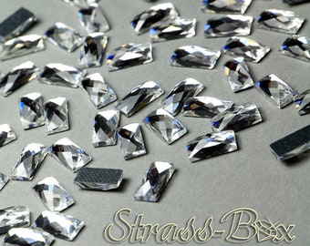 May include: A large quantity of clear, faceted, rectangular rhinestones. The rhinestones are scattered on a gray surface.