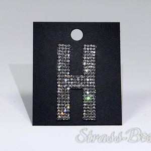 Könnte beinhalten: Ein silberner Strass-Buchstabe H auf schwarzem Hintergrund. Der Buchstabe besteht aus kleinen, funkelnden Strasssteinen.