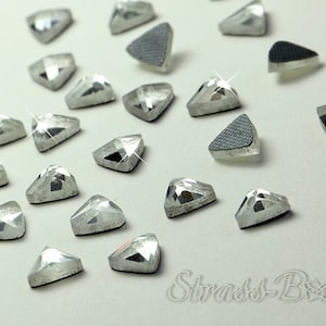 May include: A close-up of a group of silver triangle-shaped rhinestones. The rhinestones are scattered on a white surface.