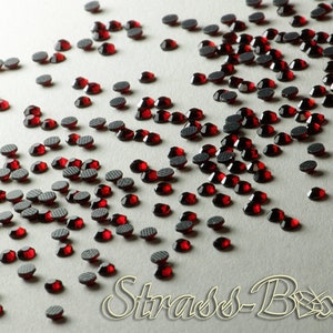 Könnte beinhalten: Eine Streuung kleiner, runder, roter Strasssteine mit flachem Rücken. Die Strasssteine sind in einem lockeren Muster auf einer weißen Oberfläche angeordnet.