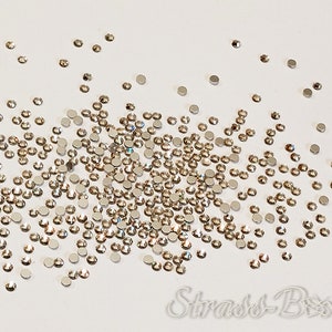 May include: A scattering of clear glass rhinestones on a white surface. The rhinestones are small and have a flat back.