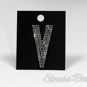 May include: A silver letter V made of rhinestones on a black card with the text "Strass-Box".