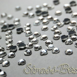 Könnte beinhalten: Silberne herzförmige Strasssteine, die auf einer grauen Oberfläche verstreut sind. Die Strasssteine haben ein facettierten Design und sind ideal zum Basteln und Dekorieren.