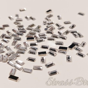 Könnte beinhalten: Eine Streuung von rechteckigen silbernen Strasssteinen auf einer weißen Oberfläche.