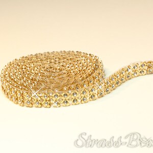 Op de afbeelding: Een opgerolde rol goudkleurige decoratieve sierband met een bloemmotief en kleine, heldere strass-steentjes. De sierband is flexibel en lijkt van een metaalachtig materiaal te zijn gemaakt. De tekst "Strass-Box" is rechtsonder zichtbaar.