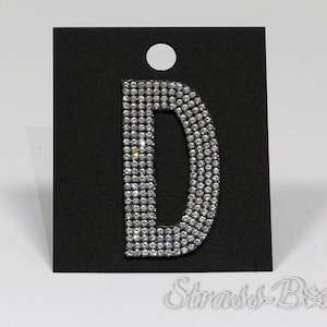 Könnte beinhalten: Ein silberner Strass-Buchstabe D auf schwarzem Hintergrund. Der Buchstabe besteht aus kleinen, runden Strasssteinen, die eng beieinander liegen. Der Buchstabe ist etwa 5 cm hoch.