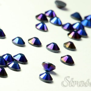 May include: A scattering of iridescent blue rhinestones on a white surface. The rhinestones have a dark blue base with a rainbow sheen.