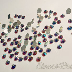 May include: A scattering of iridescent clear rhinestones on a white background. The rhinestones are various sizes and shapes, with some having a silver backing.