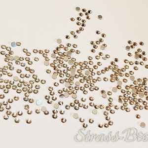 May include: A collection of scattered, circular, light brown rhinestones with a metallic backing. The rhinestones are of different sizes and are spread across a white surface. The image is well-lit, showing the reflective quality of the rhinestones. The text "Strass-Bxx" is visible.