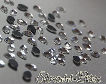 May include: A close-up of a variety of clear glass rhinestones, some with a teardrop shape, others with a round shape, and some with a more complex shape. The rhinestones are scattered on a gray surface.