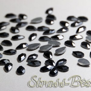 May include: Black teardrop-shaped rhinestones scattered on a white surface. The rhinestones are arranged in a random pattern.