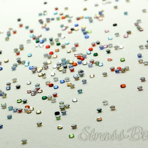 Könnte beinhalten: Sortiment aus bunten Strasssteinen, die auf einer weißen Oberfläche verstreut sind. Die Strasssteine sind klein und rund, in verschiedenen Farben, darunter Rot, Blau, Grün und Klar.