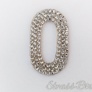 Könnte beinhalten: Ovaler Strasssteinbesatz mit klarem Rand. Die klaren Strasssteine sind dicht gepackt und erzeugen einen funkelnden Effekt. Der Besatz ist vor einem weißen Hintergrund platziert. Der Text "Strass-Bixx" befindet sich unten.