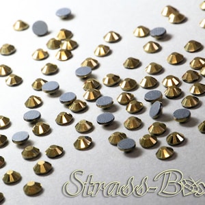 May include: A close-up of a large quantity of gold-colored rhinestones scattered on a white surface. The rhinestones have a faceted design and are used for crafting and decorating.