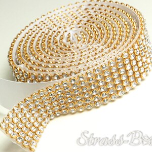 Puede incluir: Un rollo de cinta adhesiva dorada con diamantes de imitación transparentes. La cinta está enrollada en un soporte blanco.