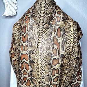 Peut inclure: Une peau de cuir de serpent, présentant un motif d'écailles marron et beige avec des marques brun foncé. La peau est drapée sur une surface blanche, mettant en valeur la texture naturelle et les variations de couleur.