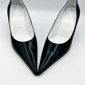 May include: A pair of black leather court shoes with pointed toes and stiletto heels. The shoes have a white interior. The brand name is visible inside the shoe. The shoes are set against a white background.
