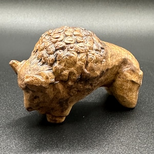 May include: A brown ceramic bison figurine with a textured surface. The bison is standing with its head lowered and its horns visible.