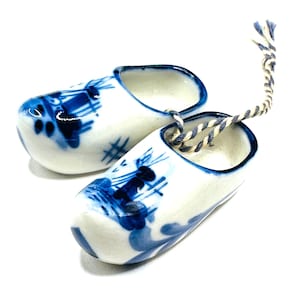 Könnte beinhalten: Ein Paar Miniatur-Holzschuhe in Weiß mit blauen Blumen- und Windmühlenmustern. Die Holzschuhe haben einen blauen Rand und sind durch eine gedrehte blau-weiße Kordel verbunden. Diese dekorativen Holzschuhe repräsentieren die niederländische Kultur.