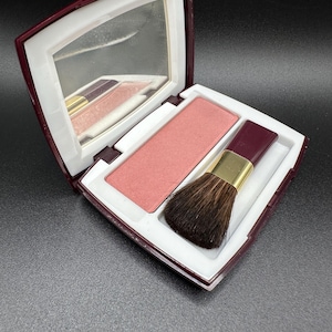 Könnte beinhalten: Geöffnete Rouge-Kompaktverpackung in Burgunderrot und Weiß mit Spiegel, einem hellrosa Rouge-Pfännchen und einem Make-up-Pinsel. Der Pinsel hat einen dunkelbraunen Kopf und einen goldenen Griff. Auf dunkler Oberfläche.