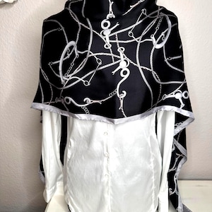 May include: A black and white patterned scarf draped over a white button-down shirt. The scarf features a repeating design of chains and equestrian-style bits. The scarf has a silver trim around the edges.