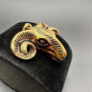 May include: A gold-colored ram head pendant with detailed features, including curled horns and textured fur. The pendant is displayed on a black velvet surface, showcasing its intricate design. This jewelry piece is likely a necklace charm or a similar accessory.