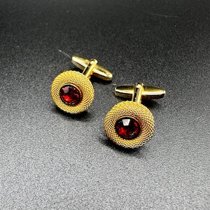 Könnte beinhalten: Ein Paar goldfarbene Manschettenknöpfe. Jeder Manschettenknopf hat eine gewebte goldene Umrandung und einen zentralen, oval geformten, tiefroten Edelstein. Die Manschettenknöpfe haben einen Klappbügel.
