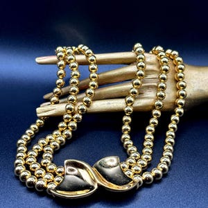 Puede incluir: Un collar dorado con tres hilos de pequeñas cuentas redondas. El collar presenta un cierre decorativo con un diseño en forma de lazo. La joya se exhibe sobre un fondo azul oscuro.