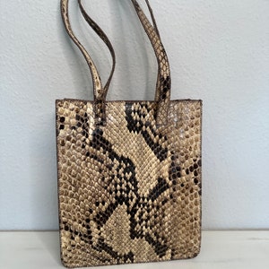 Könnte beinhalten: Eine kleine, rechteckige Handtasche aus brauner und beiger Schlangenhaut. Die Tasche hat zwei obere Henkel und einen Reißverschluss.