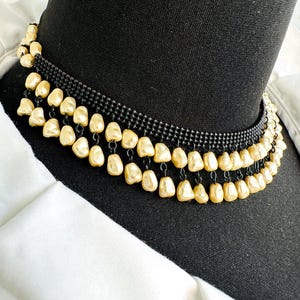 May include: A black beaded choker necklace with three rows of dangling, teardrop-shaped, cream-colored faux pearls. The necklace is displayed on a black mannequin neck, against a white shirt.