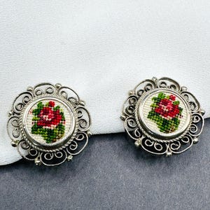 Könnte beinhalten: Paar Vintage-Ohrringe in Silberoptik. Jeder Ohrring hat ein zentrales gesticktes Design einer roten Rose mit grünen Blättern auf weißem Hintergrund, umrahmt von einem verzierten Filigranrand. Die Ohrringe sind vor einem weißen und grauen Hintergrund platziert.